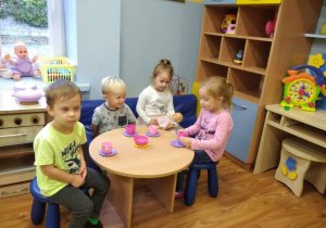 Czworo dzieci bawi się w kąciku lalek, siedzą przy okrągłym stoliku i naśladują picie herbaty z kubeczków.